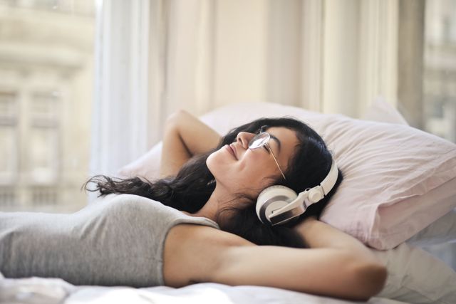 Žena u sivoj majici leži na krevetu i sluša muziku sa slušalicama
